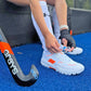 Person tying shoelaces on white sports shoes with orange accents, standing on a blue surface with a Grays hockey stick.
