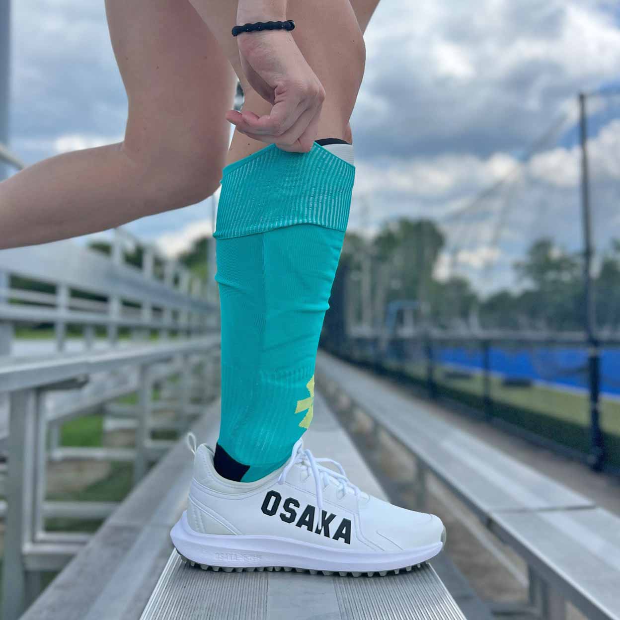 Person wearing turquoise shin guard socks and white sneakers with 'OSAKA' branding on a sports field.