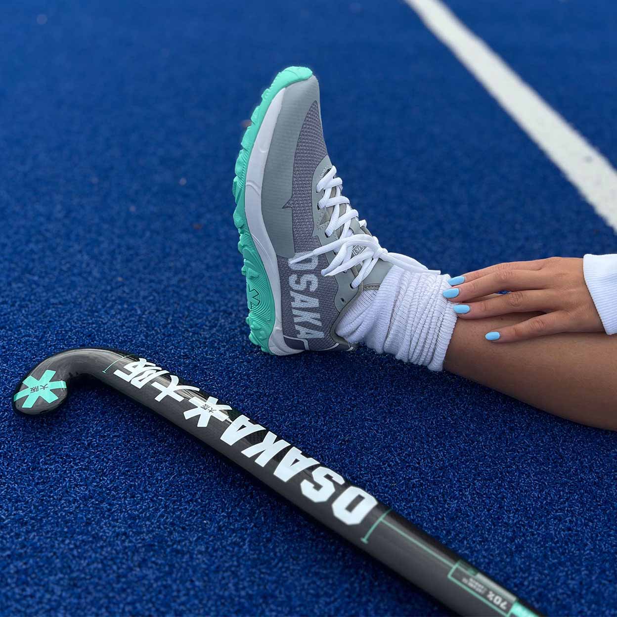 A foot wearing an Osaka Kai Field Hockey Turf Shoe next to a stick on a blue turf.