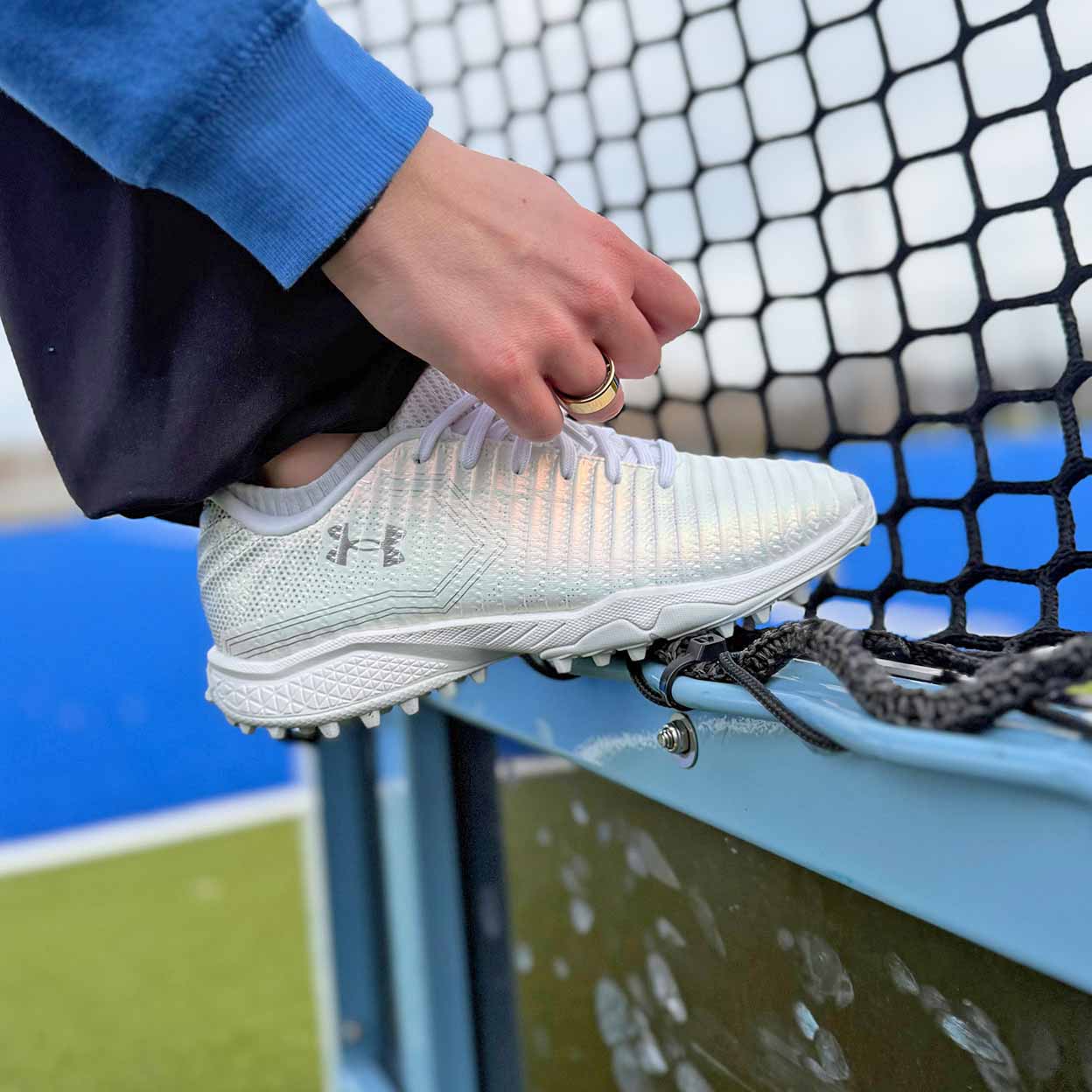 White sports shoe with cleats being worn, held by a person against a sports net background.