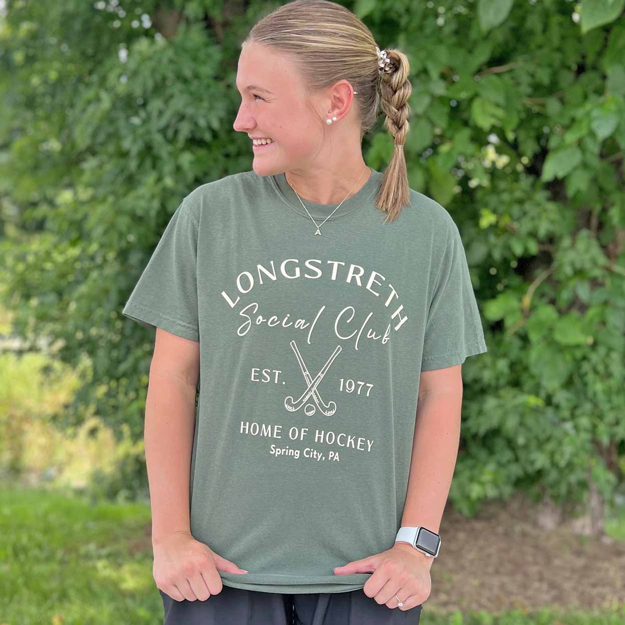 Person wearing a green t-shirt with 'Longstreth Social Club' text and graphics outdoors.