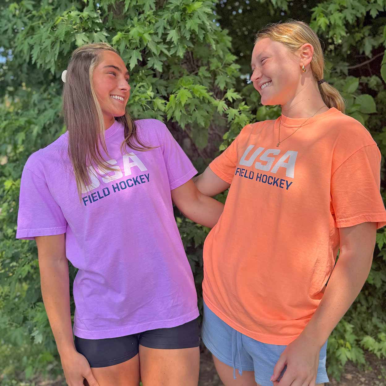 Two young women standing outdoors in front of green foliage, each wearing a short-sleeve graphic T-shirt. The shirt on the left is light purple with white and navy text reading 'USA FIELD HOCKEY,' and the shirt on the right is coral orange with white and navy text reading 'USA FIELD HOCKEY.
