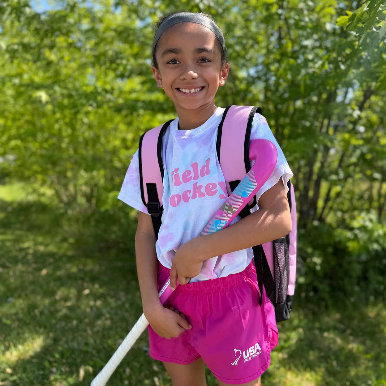 A smiling young girl outdoors in a grassy, wooded setting, wearing a white t-shirt with pink 'field hockey' text, bright pink athletic shorts with a white USA Hockey logo, and a pink backpack while holding a hockey stick.