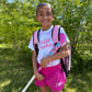 A smiling young girl outdoors in a grassy, wooded setting, wearing a white t-shirt with pink 'field hockey' text, bright pink athletic shorts with a white USA Hockey logo, and a pink backpack while holding a hockey stick.