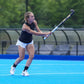 A female field hockey player in a black sleeveless top and white shorts swings a field hockey stick on a bright blue court.