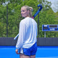 A female athlete on a blue court holding a Voodoo brand field hockey stick, wearing a white long-sleeve shirt and blue athletic shorts, looking back over her shoulder in an outdoor setting with trees and a scoreboard in the background.