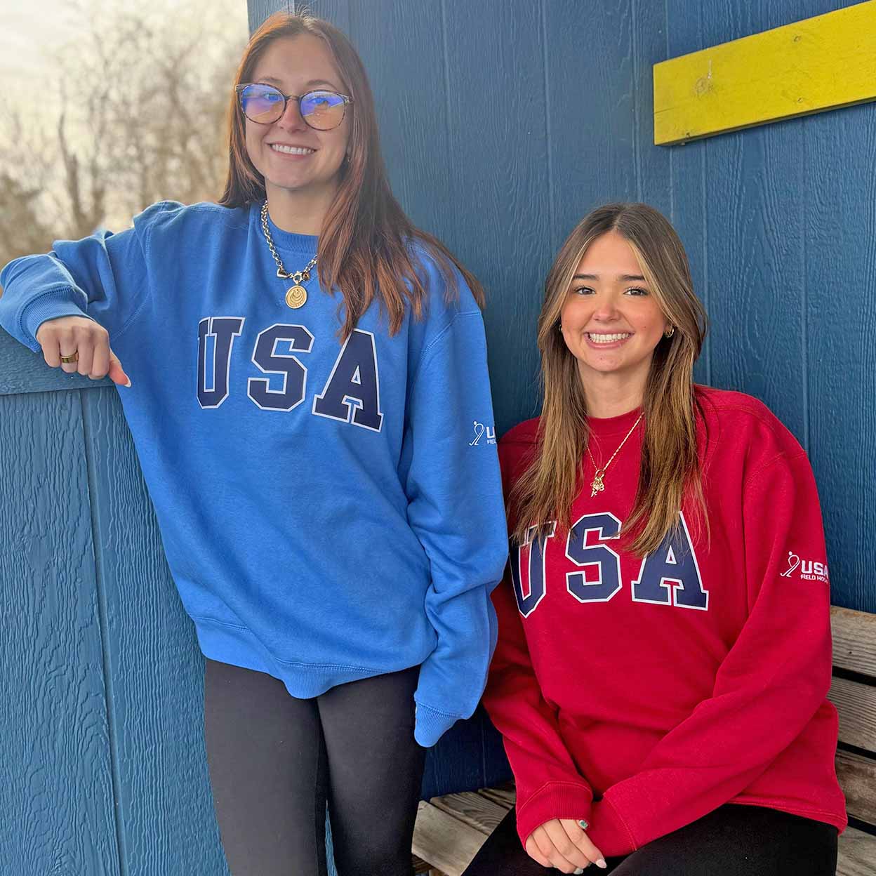 Two women wearing blue and red 'USA' sweatshirts standing against a blue wall.