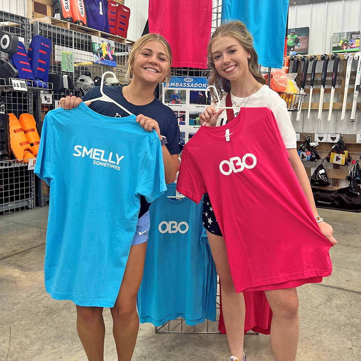 Two women holding t-shirts with 'SMELLY SOMETIMES' and 'OBO' text in a store setting.