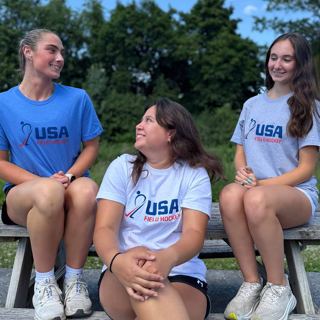 Three young female athletes sitting on a wooden picnic bench outdoors, wearing matching USA Field Hockey t-shirts in blue, white, and light gray. The shirts feature the USA Field Hockey logo in blue and red.