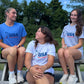 Three young female athletes sitting on a wooden picnic bench outdoors, wearing matching USA Field Hockey t-shirts in blue, white, and light gray. The shirts feature the USA Field Hockey logo in blue and red.