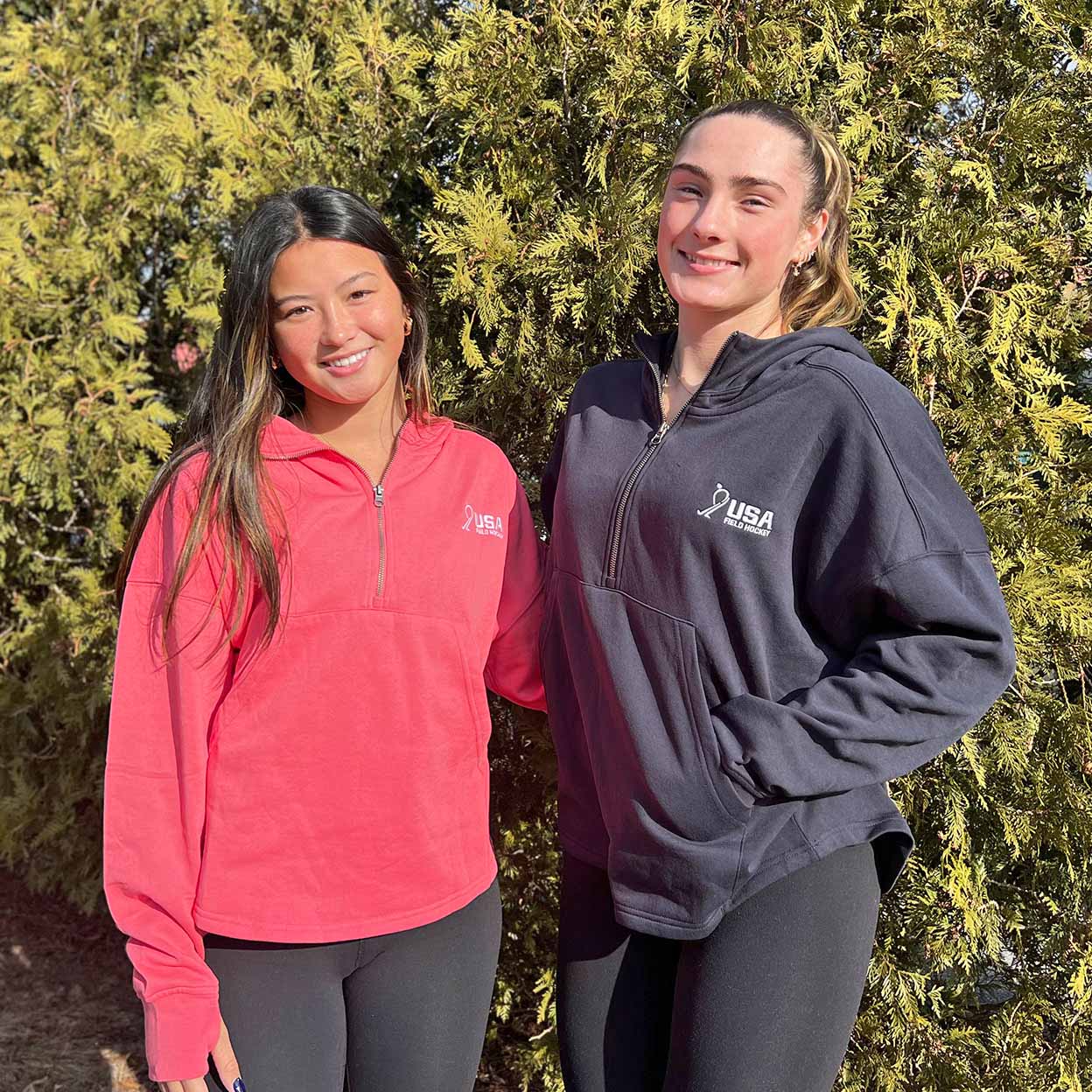 Two women wearing athletic jackets standing outdoors with greenery in the background
