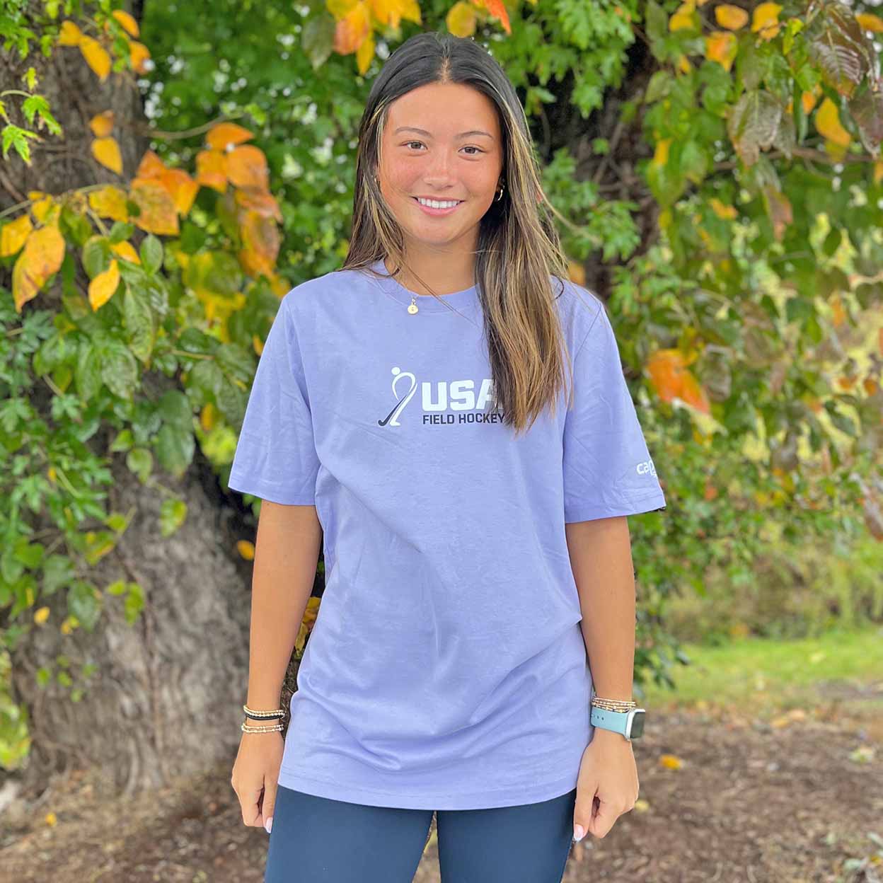 Person wearing a purple t-shirt with 'USA Field Hockey' branding against a green leafy background
