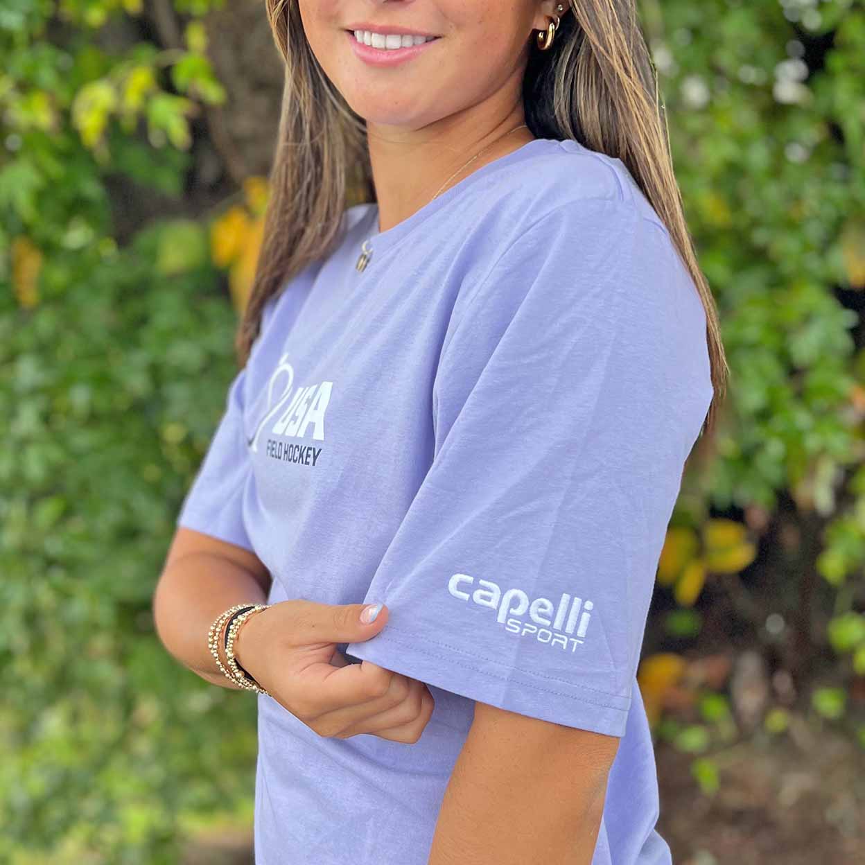 Person wearing a purple t-shirt with 'capelli sport' branding against a green leafy background