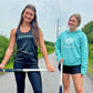 Two women standing on a road, one holding a lacrosse stick, wearing a 'Gryphon' tank top and shorts, the other in a 'Gryphon' hoodie and shorts.