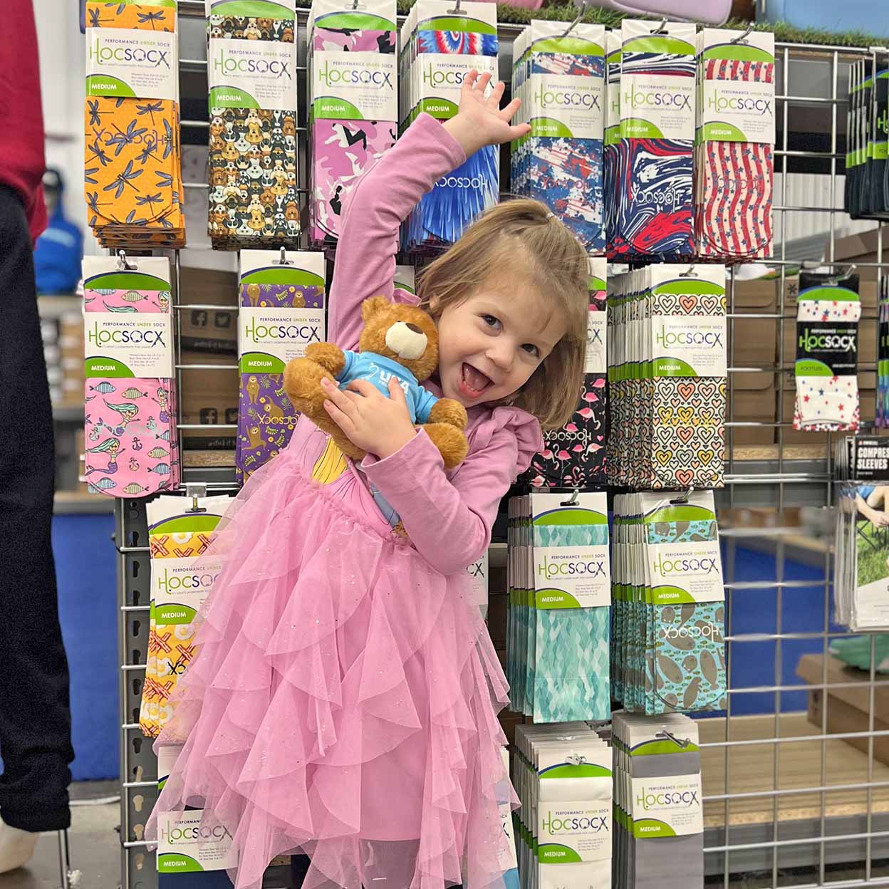 Child in a pink dress holding a teddy bear surrounded by colorful socks in a store setting