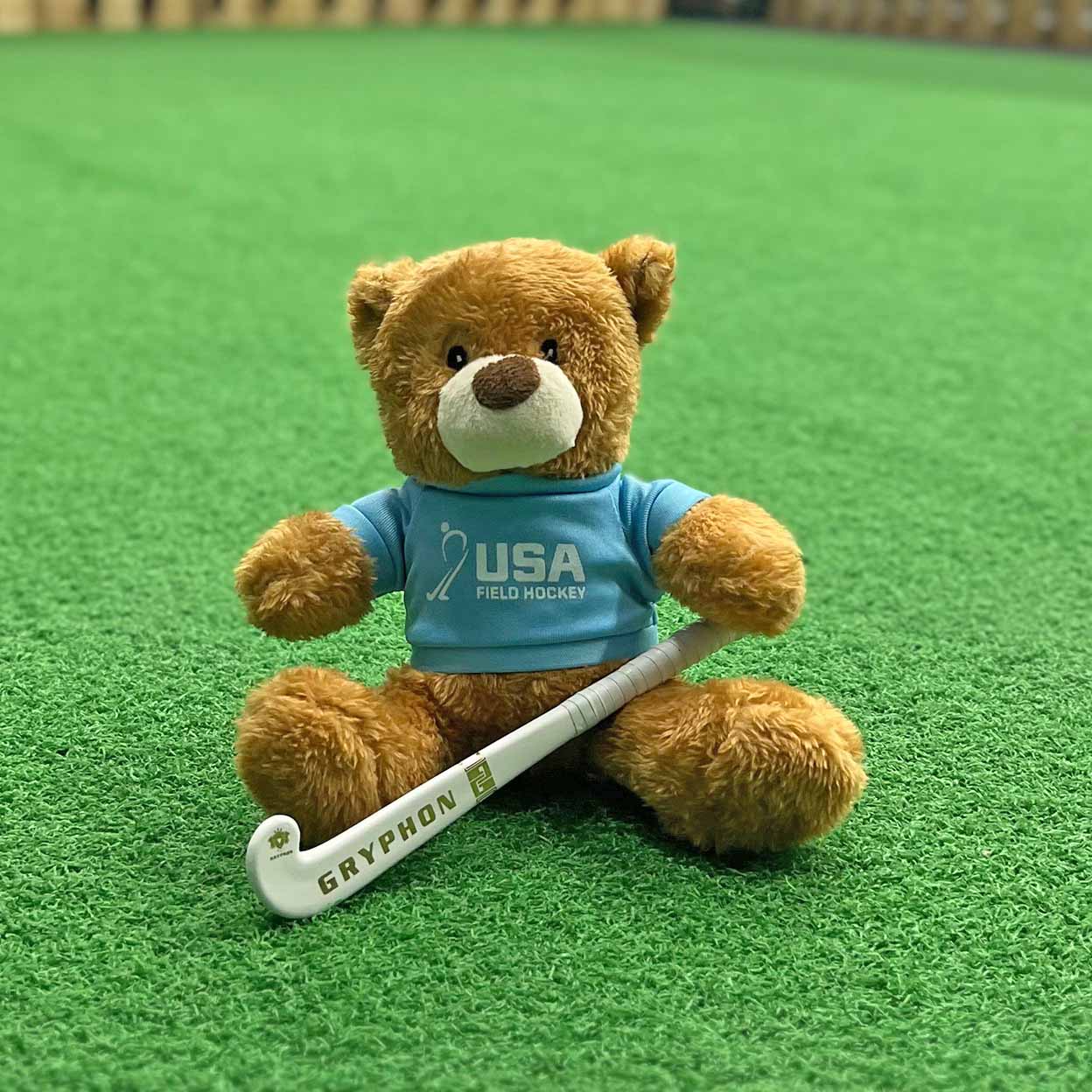 Teddy bear wearing a blue USA Field Hockey shirt with a Gryphon hockey stick on a green field background
