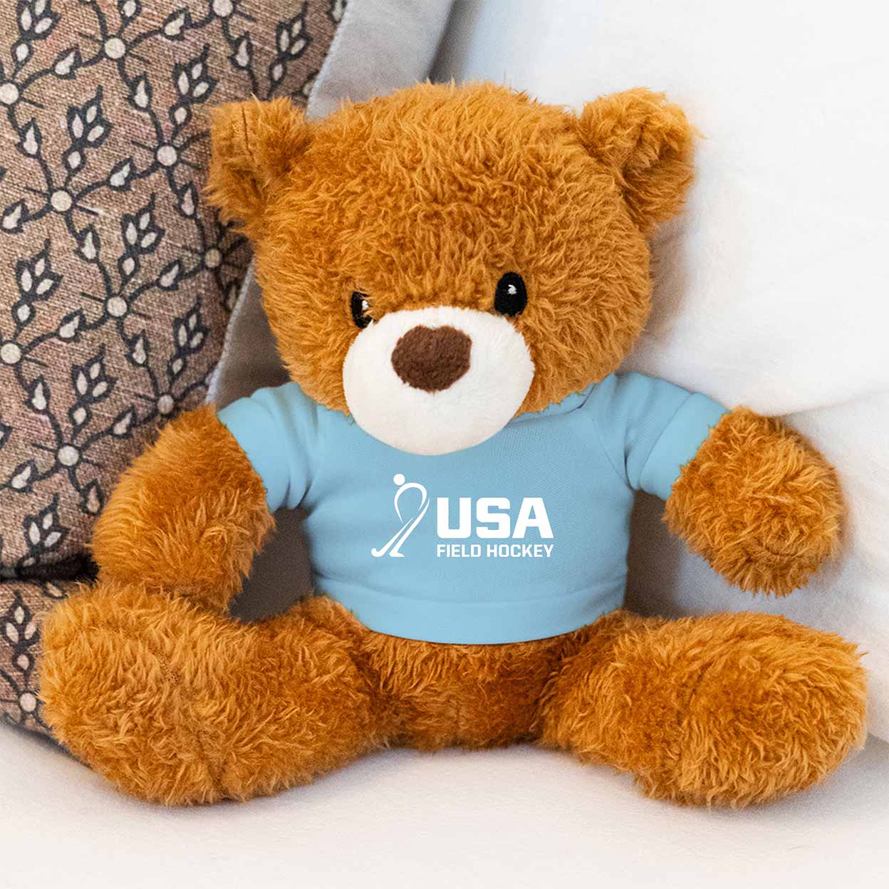 Brown teddy bear wearing a blue shirt with USA Field Hockey logo on a white background