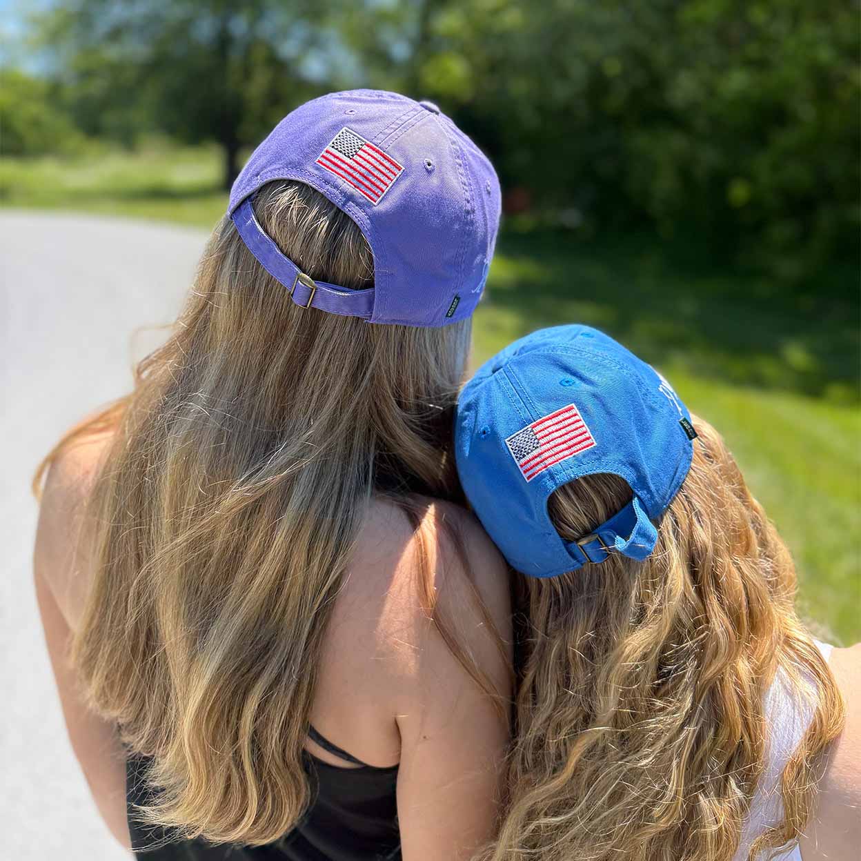Two women seen from behind outdoors, wearing baseball caps with American flag patches. One cap is lavender-purple and the other is bright blue, both showing the back with the flag patch and buckle closure.