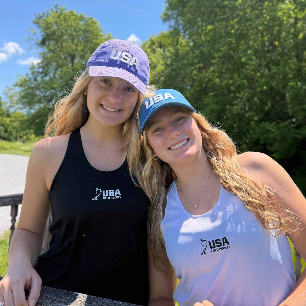 Two smiling women outdoors wearing matching USA Field Hockey athletic tank tops and baseball caps. One cap is purple and the other is blue, both with white 'USA' lettering.