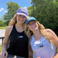 Two smiling women outdoors wearing matching USA Field Hockey athletic tank tops and baseball caps. One cap is purple and the other is blue, both with white 'USA' lettering.
