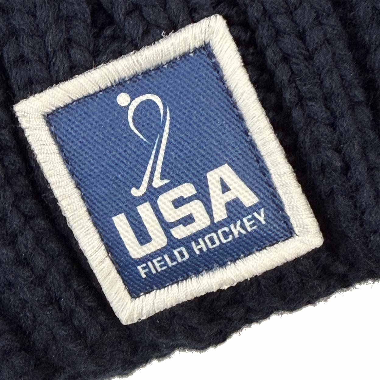 A square logo patch on a navy blue knit hat, featuring a white silhouette of a field hockey player above the text USA FIELD HOCKEY.
