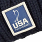 A square logo patch on a navy blue knit hat, featuring a white silhouette of a field hockey player above the text USA FIELD HOCKEY.