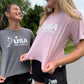 Two young women outdoors wearing USA Field Hockey athletic t-shirts, one in heather gray and one in dusty pink, both with black shorts.