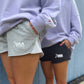 Two people wearing USA Field Hockey branded shorts, one in heather gray and one in black, paired with light purple sweatshirts, standing against a blue wall.