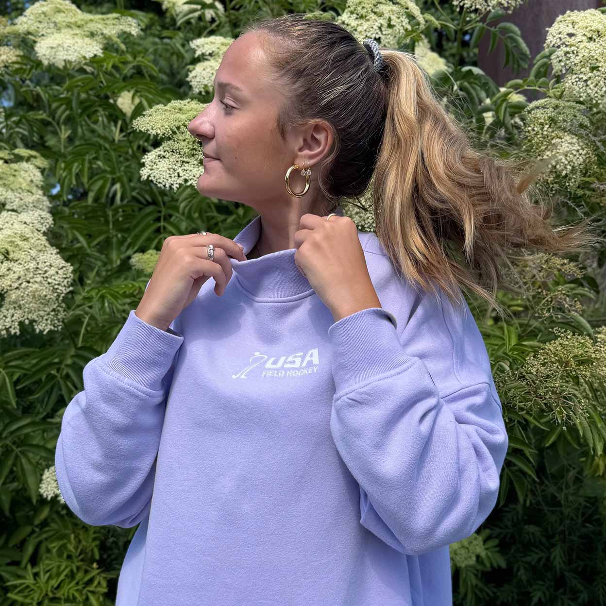 A woman wearing a light lavender mockneck sweatshirt with a white 'USA Field Hockey' logo on the chest, posing outdoors in front of green foliage and white flowers.