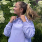 A woman wearing a light lavender mockneck sweatshirt with a white 'USA Field Hockey' logo on the chest, posing outdoors in front of green foliage and white flowers.