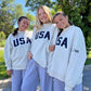 Three women standing outdoors in matching light heather gray sweatshirts with large navy USA lettering across the chest, paired with gray sweatpants.