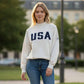 Woman wearing a white sweatshirt with 'USA' printed on it, standing outdoors.