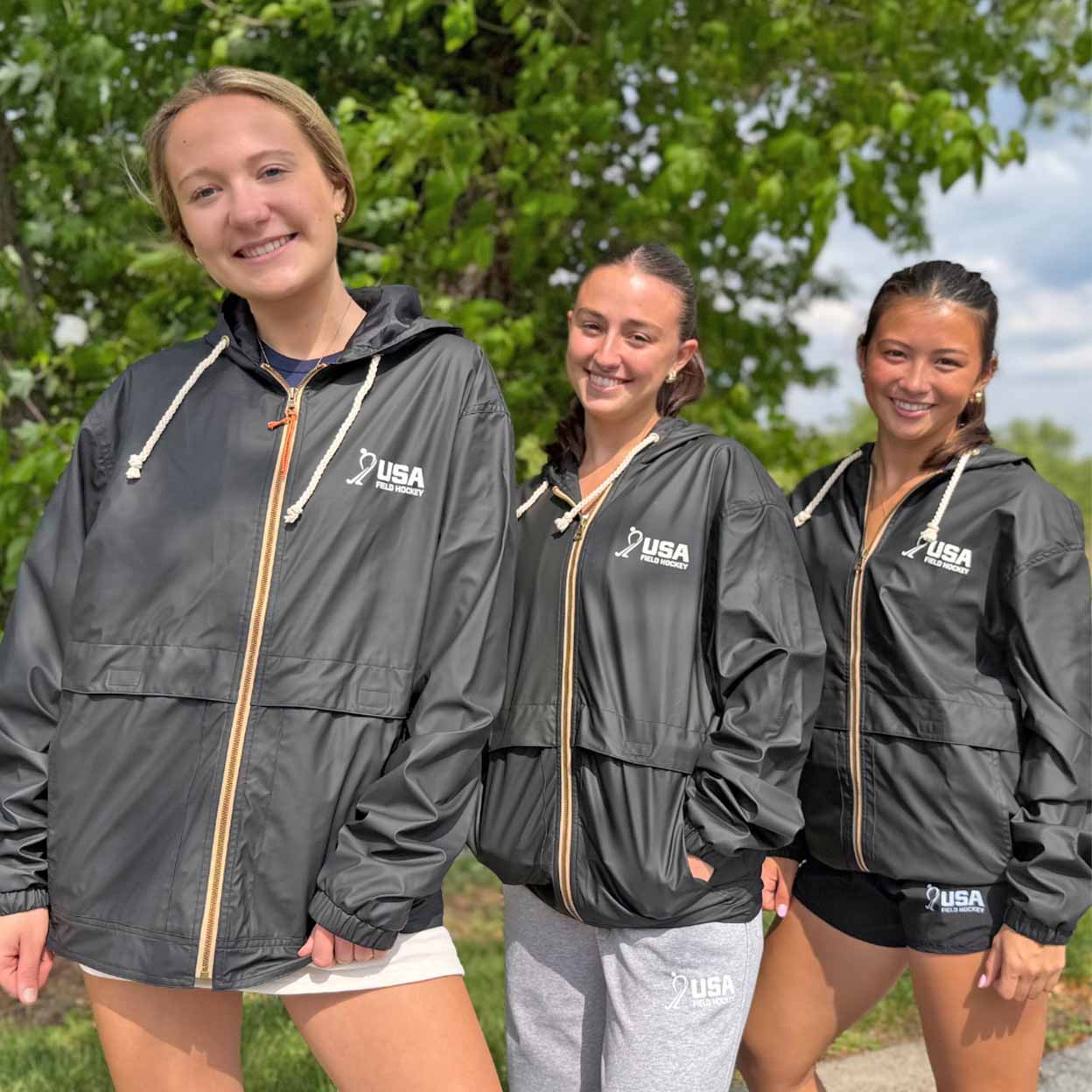 Three women standing outdoors in front of green trees, wearing matching black USA Field Hockey windbreakers with gold zippers and white USA logo on the chest.