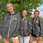 Three women standing outdoors in front of green trees, wearing matching black USA Field Hockey windbreakers with gold zippers and white USA logo on the chest.