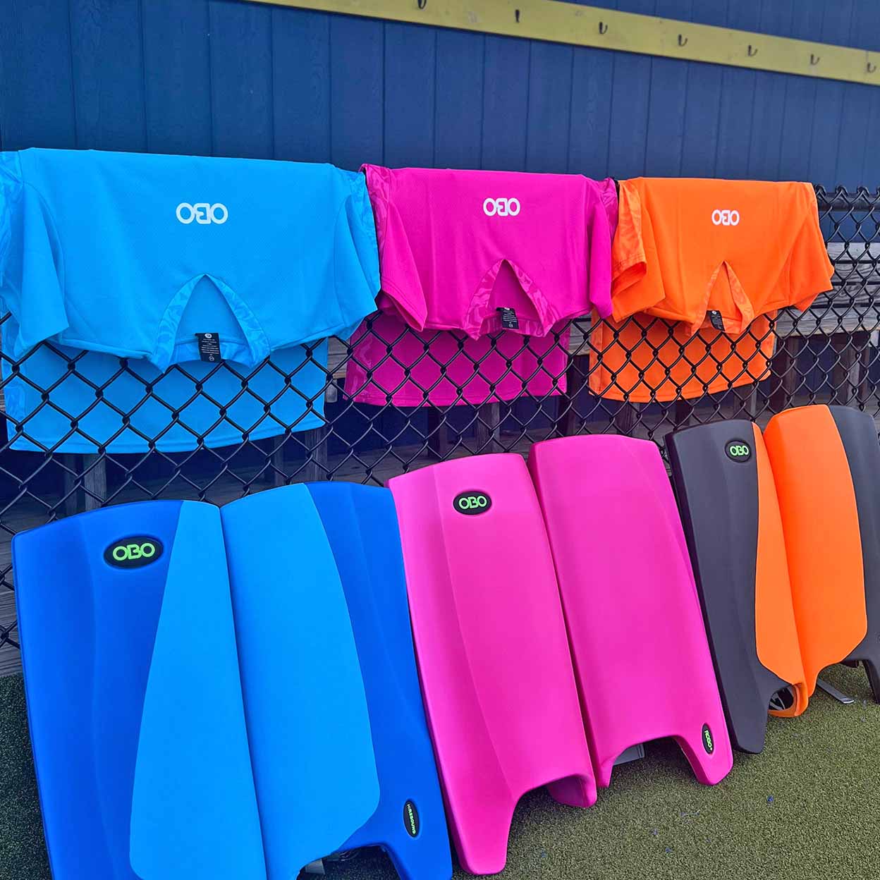 Colorful athletic jerseys and leg guards with 'OBO' branding against a chain-link fence background.