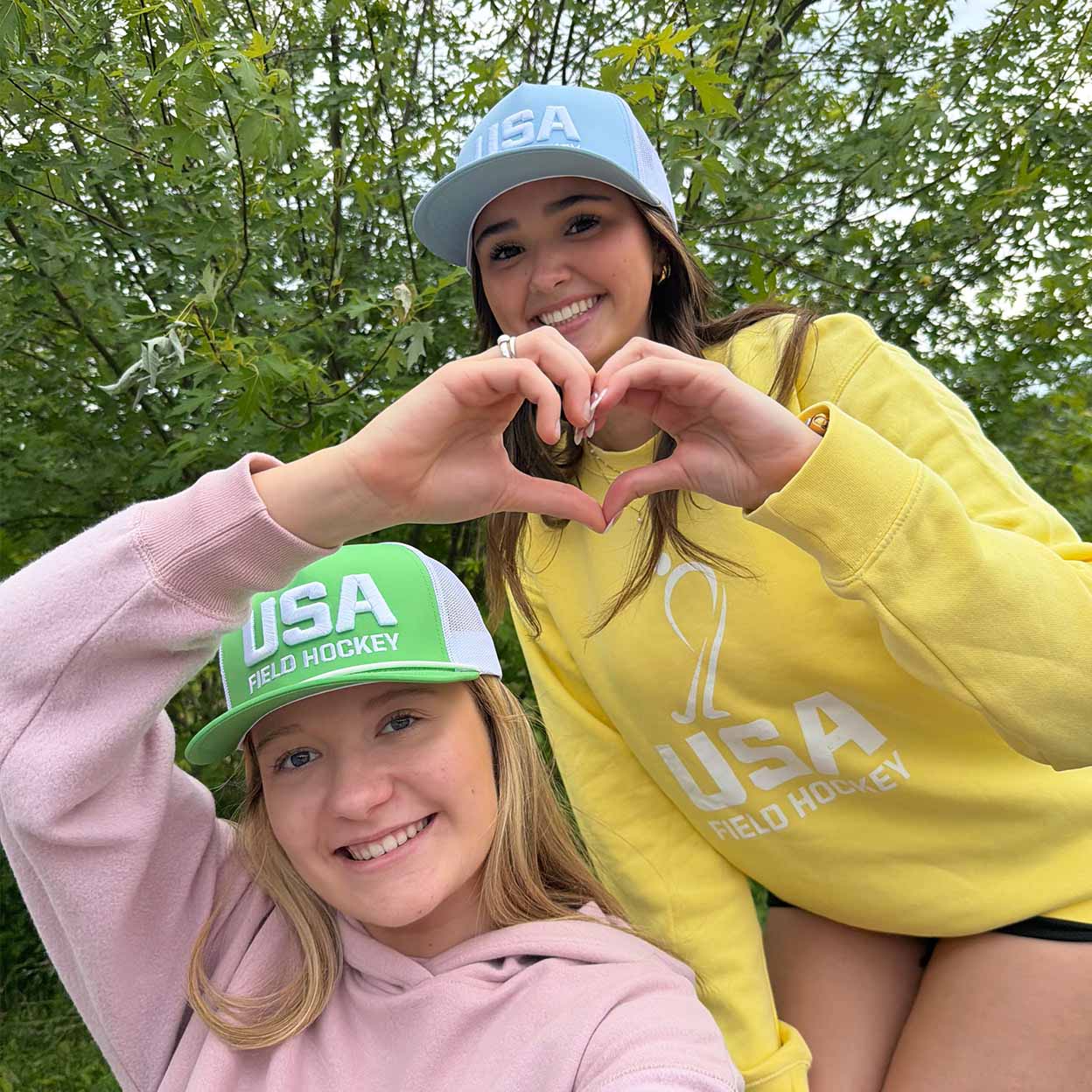 Two young women outdoors in front of leafy trees, posing together and making a heart shape with their hands. One wears a light blue USA Field Hockey cap and a yellow sweatshirt, and the other wears a green and white USA Field Hockey cap and a pink sweatshirt.