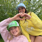 Two young women outdoors in front of leafy trees, posing together and making a heart shape with their hands. One wears a light blue USA Field Hockey cap and a yellow sweatshirt, and the other wears a green and white USA Field Hockey cap and a pink sweatshirt.