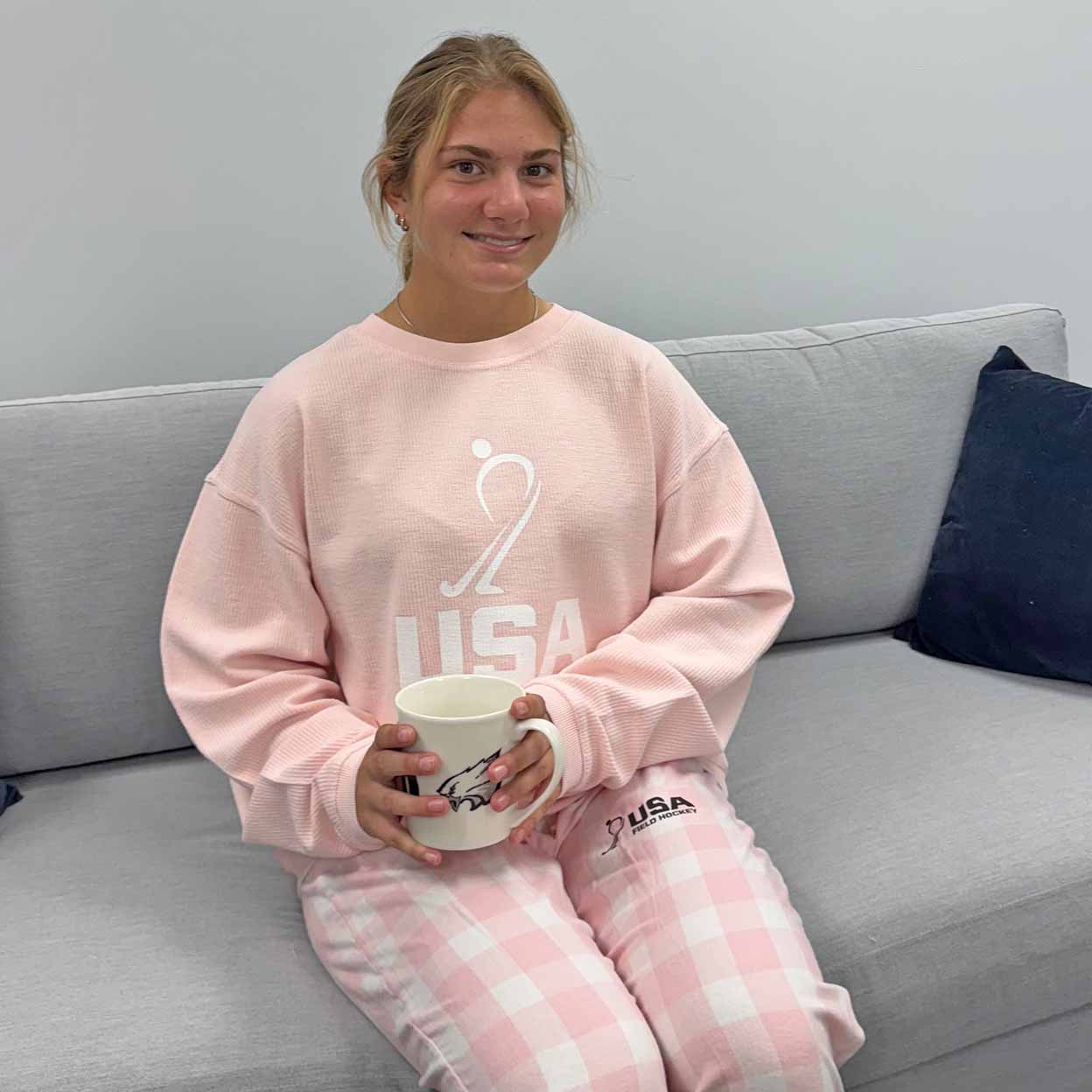 Person wearing pink 'USA' pajamas sitting on a gray couch holding a white mug.