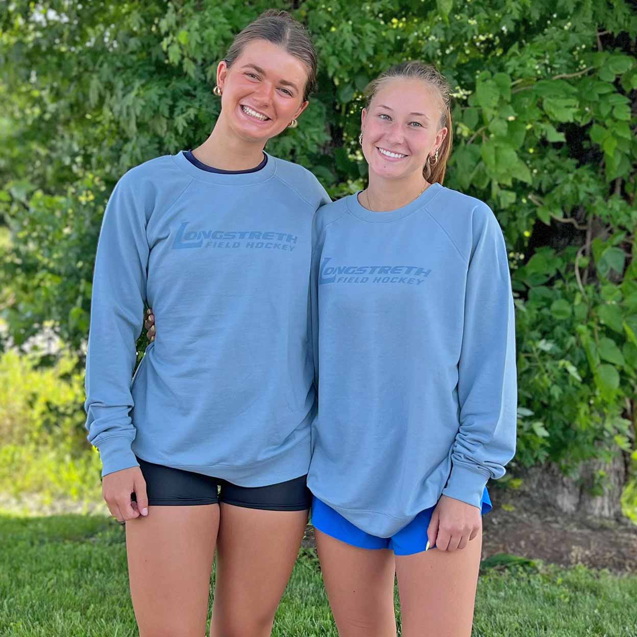 Two people wearing matching blue sweatshirts with a logo in front of green foliage.