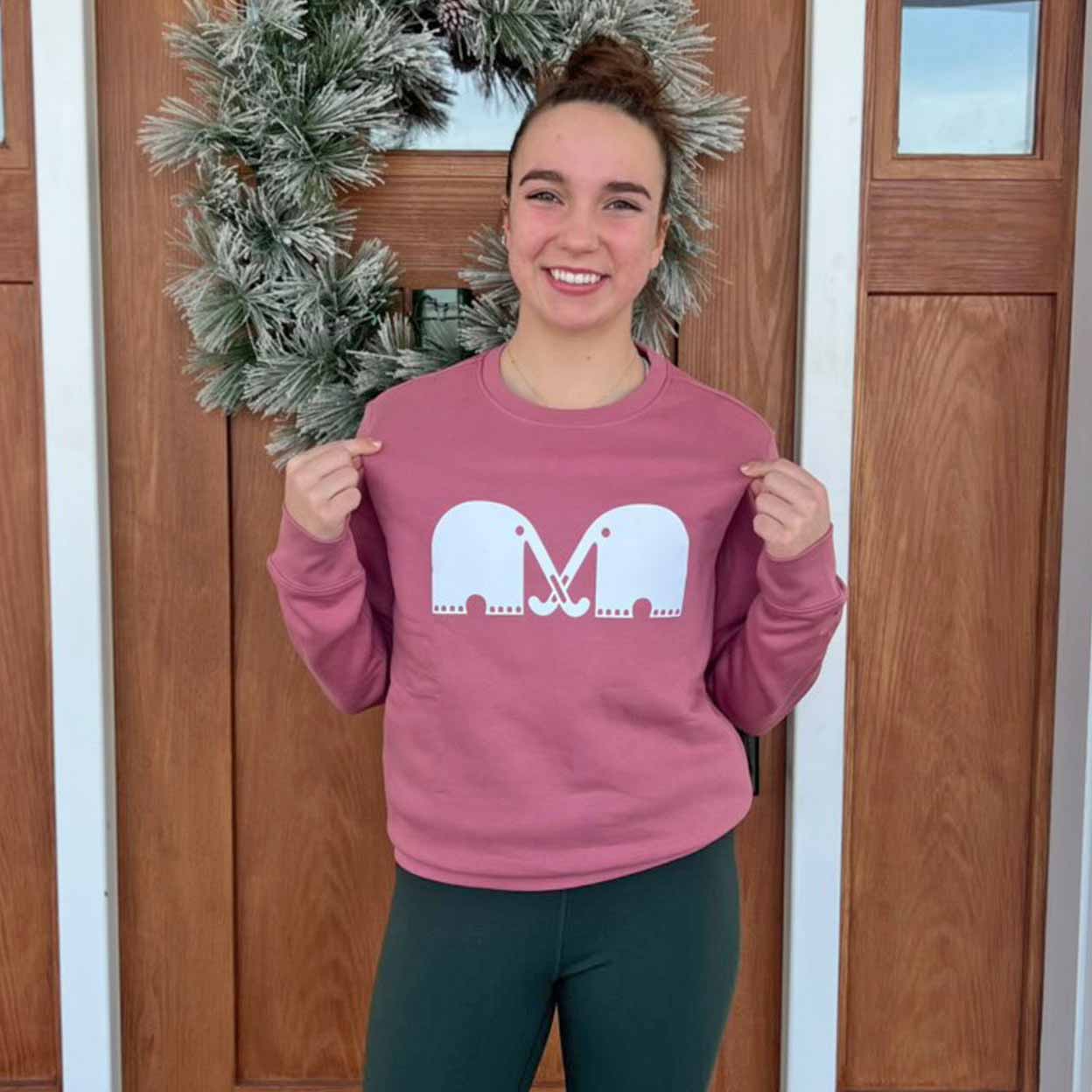 Person wearing a pink sweatshirt with a white elephant design in front of a wooden door with a wreath.