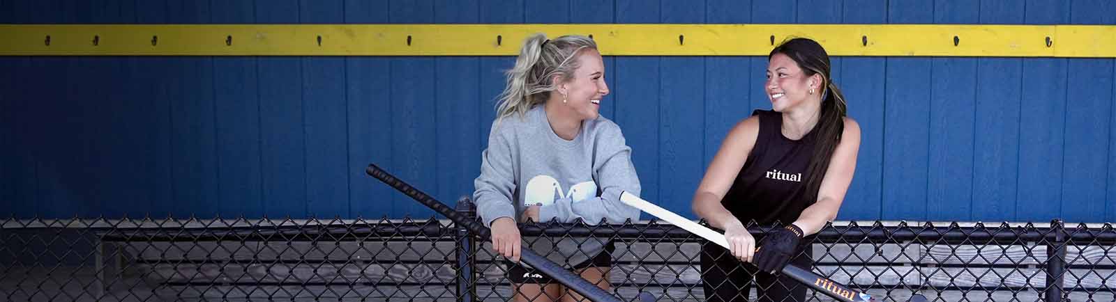 Two women holding hockey sticks in front of a blue and yellow wall.