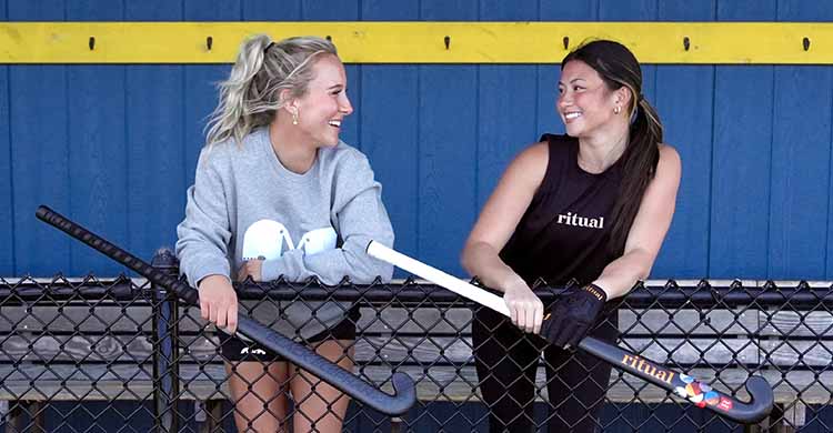 Two women holding hockey sticks in front of a blue and yellow wall.